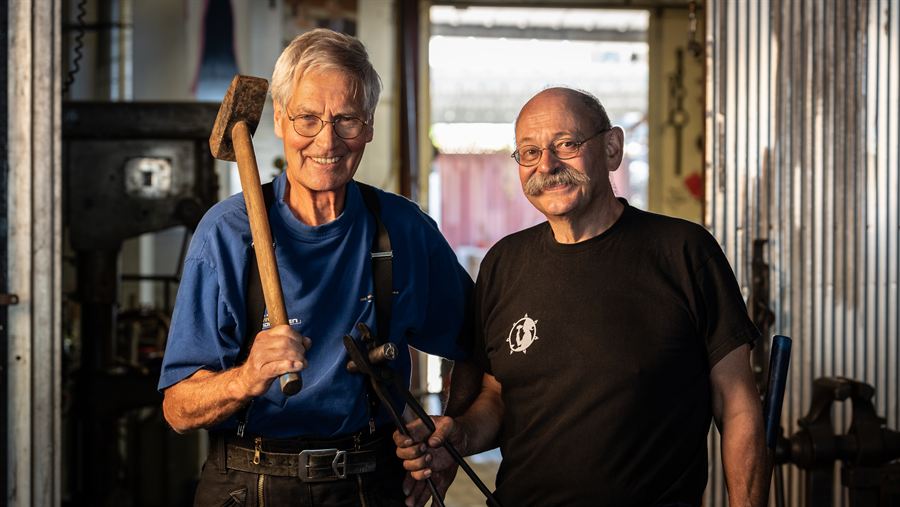 Twee smeden lachend in de werkplaats, met een voorhamer en een smeedtang